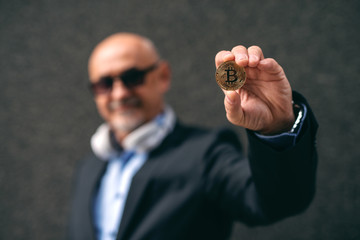 Bearded senior adult wealthy businessman holding bit-coin. Selective focus on bit-coin.