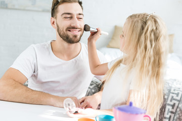 adorable little daughter applying makeup to happy bearded father at home