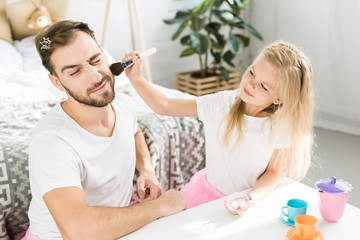 Obraz na płótnie Canvas smiling little daughter applying makeup to bearded father at home