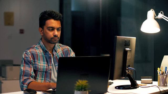 business, technology and creative people concept - young indian man with laptop computer working at dark night office