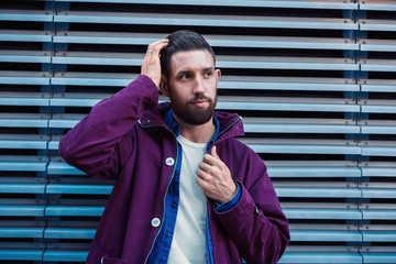 Handsome young man with hairstyle in fashionable clothes stands near iron wall and looking sideways