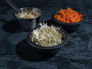 Preparation for the salad, slicing carrot cabbage in three cups, grated cheese on a dark background. Close-up.