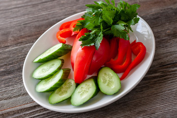 Sliced ​​fresh vegetables, sweet peppers and cucumbers on a white plate, decorated with greens, look appetizing.
