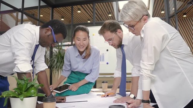 Casual office, multiracial architects working on project blueprint. Caucasian, asian, african colleagues discussing goals.