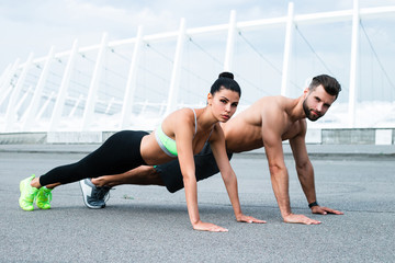 Fototapeta na wymiar Planking with us? Young beautiful couple in sportswear exercising together against industrial city view