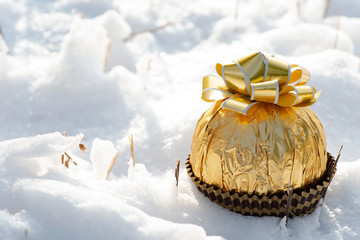 Winter holiday decoration concept: Italian chocolate ball wrapped in metallic gold foil and topped with golden ribbons, bows and frozen snow covered pine tree.