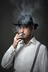 Serious old-fashioned man in hat lighting a cigar on dark background.