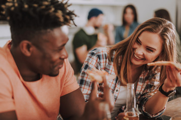 Portrait of beautiful girl holding slice of pizza and bottle of beer while looking at friend and smiling. Focus on woman