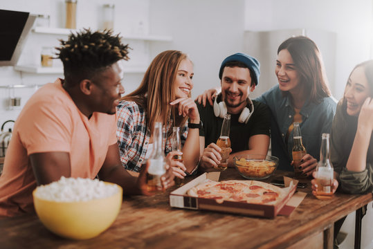 Joyful friends sitting at the table with pizza, beer, chips and popcorn. They looking at each other and smiling