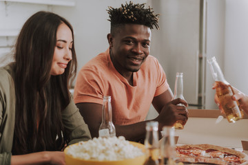 Fototapeta na wymiar Portrait of handsome afro american guy and charming girl sitting at the table with beer, popcorn and pizza. They are smiling