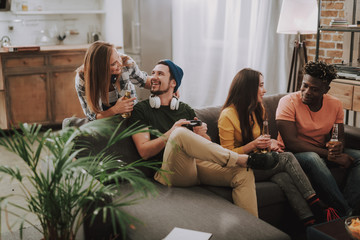 Handsome bearded man in hat using joystick while charming lady looking at him and smiling. Joyful afro american guy sitting on couch and talking with brunette lady
