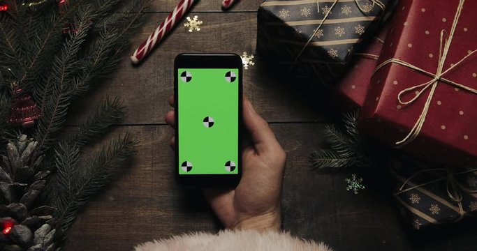 Hands of Santa Claus tapping pages on the black smartphone with green screen. Chroma key. Top view. Vertical position. Christmas New Year concept