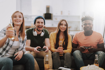 Young people sitting on couch and smiling. Guys using joysticks while girls holding bottles of beer