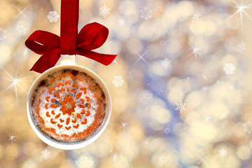 Christmas card- cup of fragrant hot coffee with white foam cinnamon snowflake made from ball, bauble hangs on red ribbon on background of defocused silver lights. Concept holidays symbol for New Year