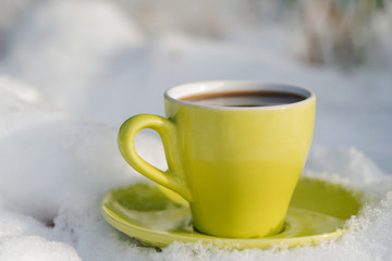 Winter holiday decoration concept: green cup of coffee with saucer on frozen snow covered wood log in forest preserve park.