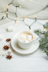 Obraz na płótnie Canvas Coffee with milk, latte with cinnamon sticks and anise stars with white chocolate and marshmallow, with a branch of a Christmas tree on a bright white background, with lights from a garland.