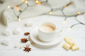 Obraz na płótnie Canvas Coffee with milk, latte with cinnamon sticks and anise stars with white chocolate and marshmallow, on a light white background, with lights from a garland.