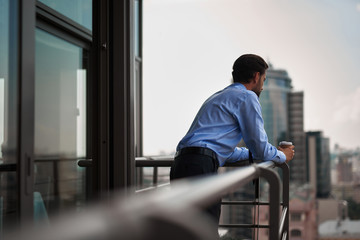 Corporate time-out culture. Back side portrait of young businessman standing on office balcony with cup of coffee and looking at city view - Powered by Adobe