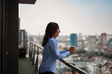 Corporate time-out. Waist up side on portrait of young smiling office woman standing on balcony with cup of coffee. Copy space on right