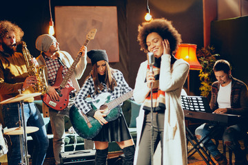 Close up of mixed race woman singing. In background band playing instruments. Home studio interior.