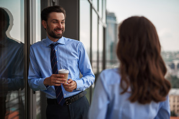 Friendly corporate culture. Waist up selective focus on young smiling man standing with woman on office balcony while drinking coffee