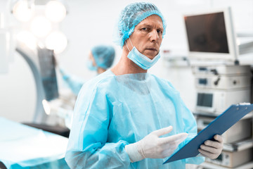 Thoughtful doctor wearing blue medical uniform and frowning suspiciously while looking and holding his clipboard