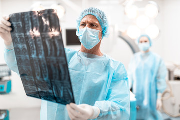 Concentrated professional doctor frowning and thoughtfully looking at the x-ray results in his hand while his colleague standing on the background