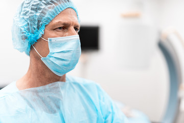 Calm professional doctor wearing medical hat and mask with his blue uniform and frowning while looking into the distance