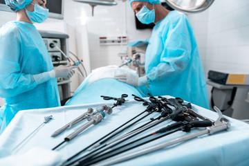 Sterile table with laparoscopic scissors, graspers, trocars with cannulas and clip applicator. Surgeon performing surgical operation while nurse looking at patient on blurred background