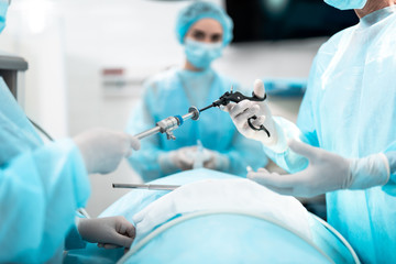 Close up of male and female hands holding laparoscopic grasper with trocar. Female medical worker holding oxygen mask on patient face on blurred background