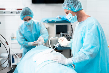 Naklejka na ściany i meble Side view portrait of middle aged doctor in protective mask and sterile gown performing surgical operation in modern operating room. Female assistant on blurred background