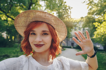 Beautiful and stylish red haired girl taking a selfie with her smartphone outdoors, authentic...