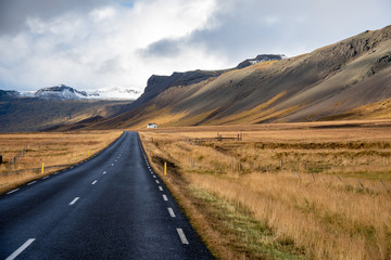 Road Running at the Foot of Towering Mountains in Iceland on a Cloudy Autumn Day. A Farm is Visible on the Right Side of the Road.