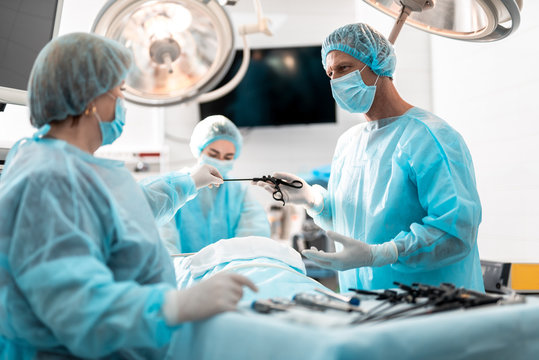 Side view portrait of surgeon in protective mask taking laparoscopic scissors while looking at assistant with serious expression