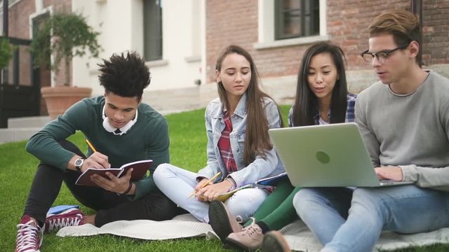 Charismatic american four person talking, sitting along college campus. Multiracial 20s friends in smiling discussion, writing, looking in computer. Friendly students using modern technology in