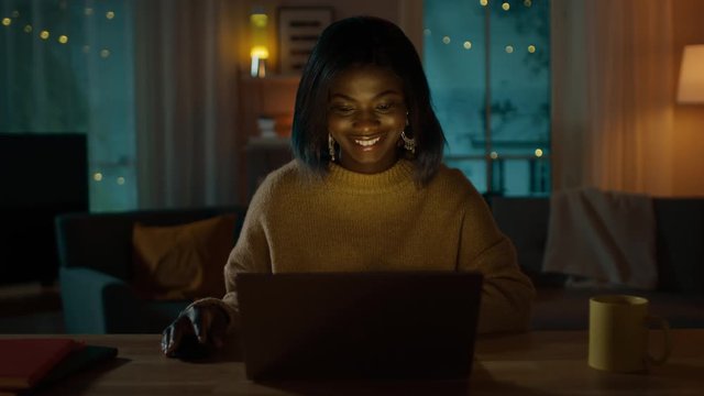 Portrait of Beautiful Smiling African American Girl Using Laptop while Sitting at Her Desk at Home. In the Evening Creative Woman Works on a Computer In Her Cozy Living Room. 