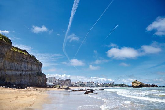 Biarritz city and its famous sand beaches. Stones, ocean waves. Atlantic coast, Basque country, France. Summer sunny day and blue sky with white airplane trails