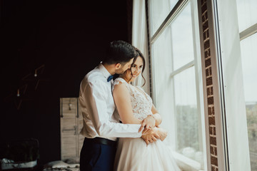 Stylish young groom gently hugs his charming brunette bride.Portrait of a young happy couple near the window on black background.