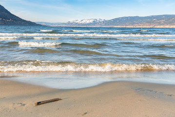 Waves crashing onto sandy beach with snow-capped mountains in distance and blue sky