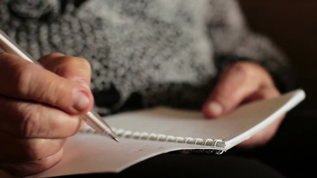 The old woman writes something in a notebook or diary. The wrinkled hand of an old woman writes something on paper