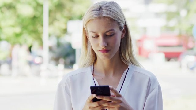 Attractive young female in white blouse browsing modern smartphone while standing on blurred background of street on sunny day
