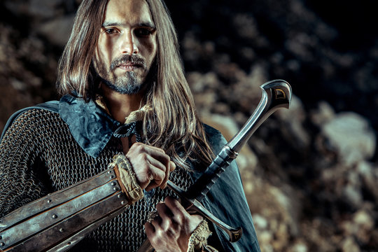 Long haired knight with the two-handed sword. Rocks on the background.