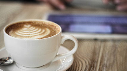 cup of coffee on a wooden table on a background of young woman works with digital tablet
