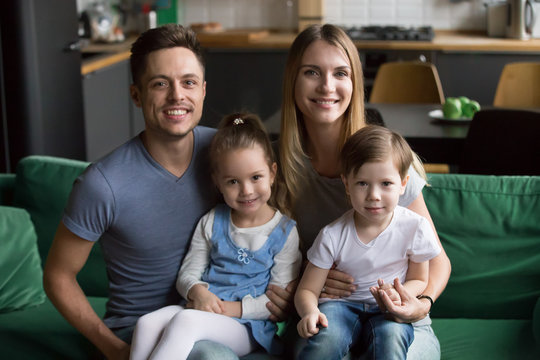 Portrait of happy smiling large family with two children, mother and father sitting on sofa at home together with son and daughter, kids sitting at parents knees, look at camera