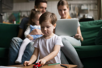 Little boy playing with toy cars while parents sitting together, playing with daughter, mother and father using laptop, watching video, son playing alone, family spending time together
