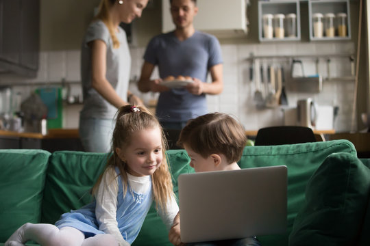 Little girl and boy, son and daughter using laptop, looking at screen, watching cartoons, playing video game while parents cooking, setting table, preparing for dinner, family spending time together