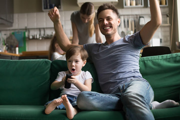 Toddler son with father watching football, baseball, basketball on TV together, excited, celebrating, rejoice at goal, victory of favorite team, mom and daughter cooking, family spending time together