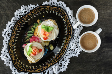 Two biscuit cakes with butter cream, decorated with flowers from the cream are on a beautiful vintage metal tray and white lace doily are next to two cup of coffee on a dark background, top view