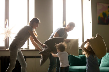 Young funny family playing pillow fight with toddler son and daughter in living room with large window at home, happy excited mother and father with children, family spending time, having fun together