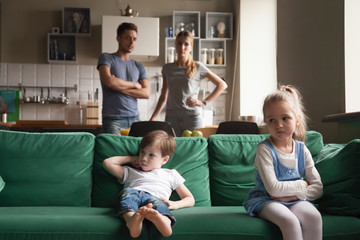 Upset offended sister, daughter sitting separately on couch with arms crossed with toddler brother...